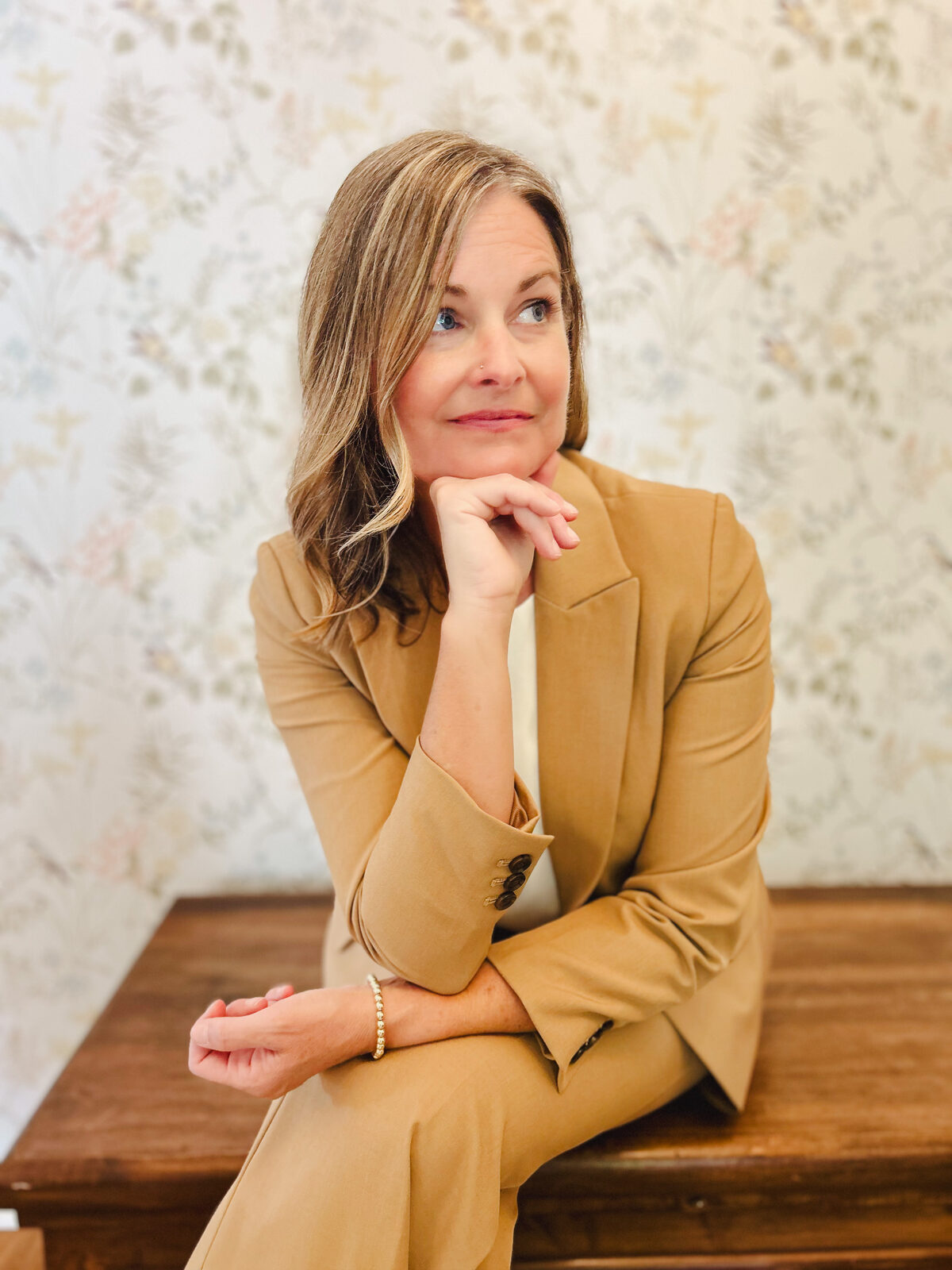 Julia in a tan suit, chin resting on her hand, seated against soft floral wallpaper