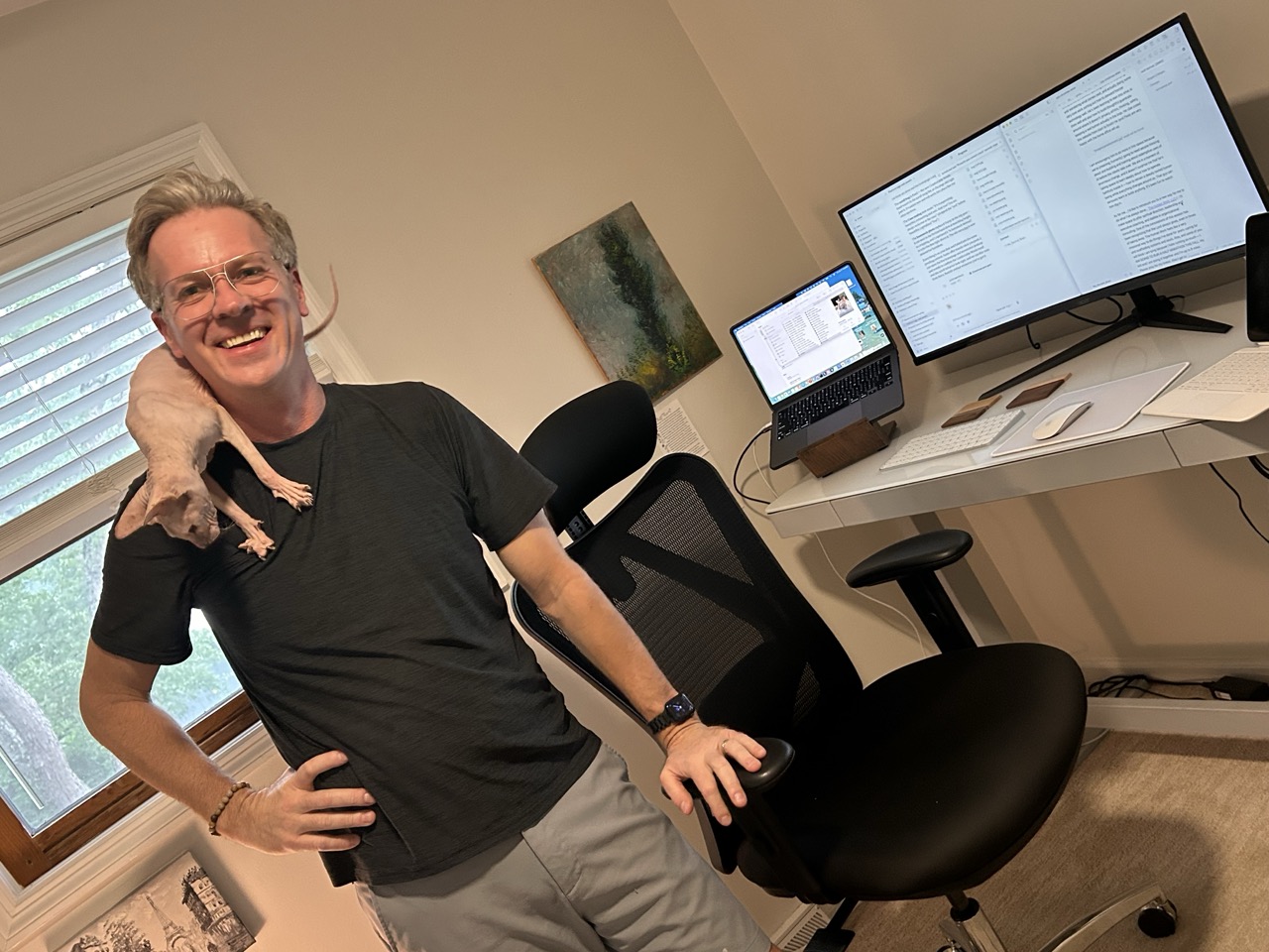 Eric grinning in his home office with Pixie draped over his shoulder, his desk and monitor behind him